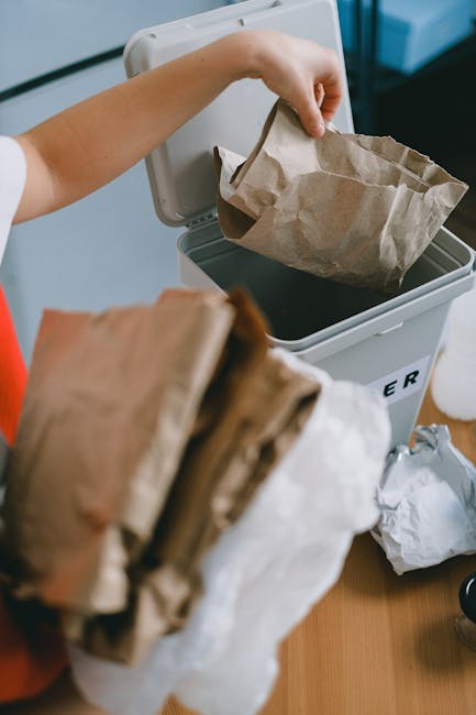 A person is seen disposing of paper waste into a white waste bin that has a partially visible label, with the lid open to reveal a brown paper bag inside. The individual is holding a crumpled paper bag in one hand while placing it into the bin, which is positioned on a wooden surface. In the foreground, another crumpled paper bag is held, appearing to contain additional waste. The environment appears to be an indoor setting, likely a kitchen or office space, with neutral lighting highlighting the textures of the paper bags and the smooth surface of the bin. The scene emphasizes the act of proper waste separation and disposal, aligning with private rubbish removal services offered by companies like rubbishremovalcrystalpalace.com, which assist with on-site clearance and alternative waste handling solutions to traditional council collections.