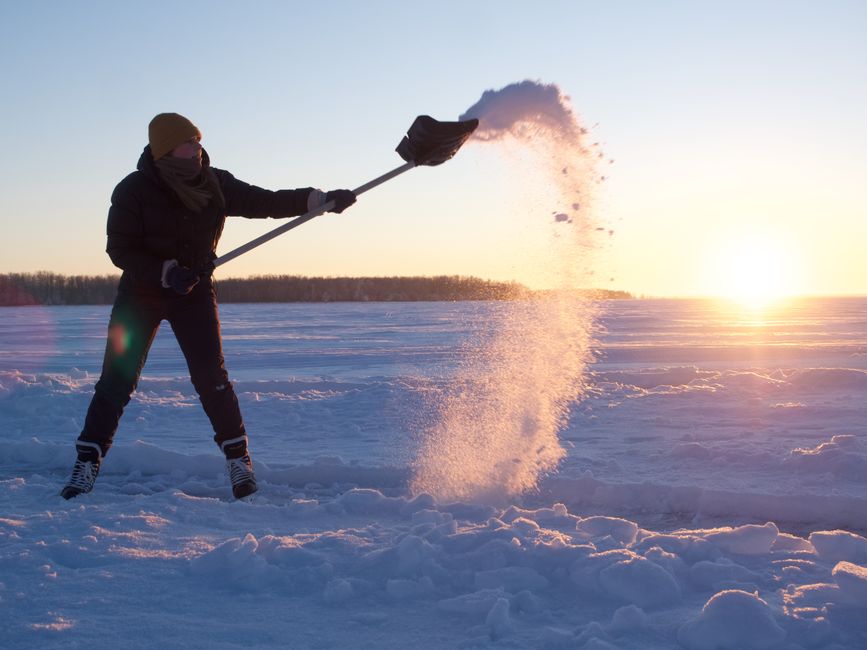 A person dressed in dark winter clothing, including a mustard-colored beanie, a black coat, gloves, and patterned winter boots, is standing on a snow-covered open field during sunset. They are holding a black snow shovel with both hands and are using it to throw a cloud of snow into the air. The snow displays small particles illuminated by the low sunlight, creating a sparkling effect. The background features a distant tree line beneath a clear sky with the sun near the horizon, casting warm, golden light across the landscape. The scene evokes a cold winter environment and showcases an outdoor activity in a natural setting, with the snow being disturbed by the person's movement, aligning with themes of snow clearing or outdoor winter work.
