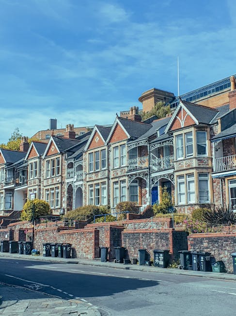 A row of Victorian-style terraced houses with detailed wrought iron balconies and decorative gables, situated on a slope above a paved street in an urban residential area. The buildings feature brick facades with white window frames and ornate trim, and some have small front gardens enclosed by brick walls with shrubbery and plants. Several black wheelie bins are lined up along the pavement at the base of the houses. The sky above is mostly clear with some light clouds, and modern commercial or office buildings are visible behind the row of houses, indicating a mixed-use neighborhood. The scene captures the everyday environment where private waste collection or rubbish removal services, such as those provided by [COMPANY_NAME], may operate for property maintenance, cleaning, or clearance projects, reflecting alternative waste handling approaches outside of municipal rubbish collection.