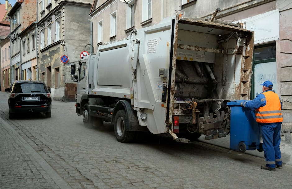 A white commercial rubbish collection truck, featuring a rusted and metal-textured rear loading compartment, is positioned on a cobblestone street alongside a row of old, multi-story buildings with weathered facades. The truck's rear door is open, revealing mechanical components used for waste compaction. A worker dressed in blue overalls and an orange high-visibility vest, wearing a blue cap, is seen standing next to a blue wheeled bin and appears to be preparing to load waste or remove the bin. In the background, a black car is parked parallel to the curb, and a no-parking sign is mounted on a pole attached to the buildings. The scene exudes a typical urban environment dedicated to scheduled waste collection or private rubbish removal, with natural daylight illuminating the scene, emphasizing the utilitarian nature of the waste management process in a residential or commercial district.