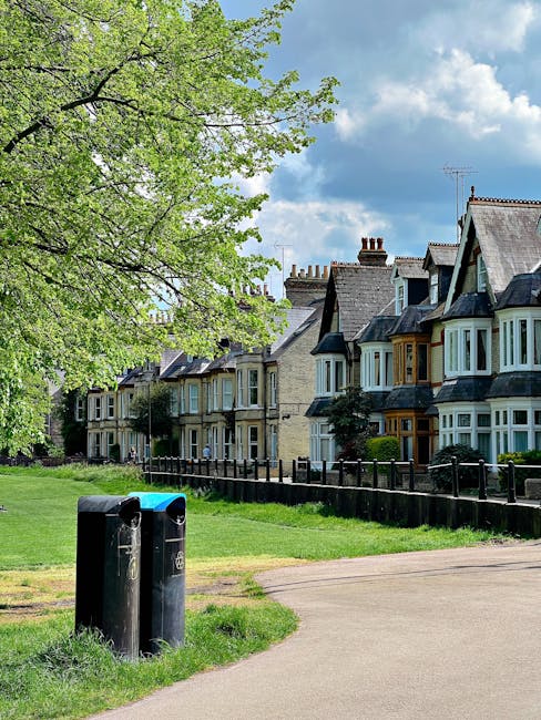 A row of Victorian-style terraced houses with detailed wrought iron balconies and decorative gables, situated on a slope above a paved street in an urban residential area. The buildings feature brick facades with white window frames and ornate trim, and some have small front gardens enclosed by brick walls with shrubbery and plants. Several black wheelie bins are lined up along the pavement at the base of the houses. The sky above is mostly clear with some light clouds, and modern commercial or office buildings are visible behind the row of houses, indicating a mixed-use neighborhood. The scene captures the everyday environment where private waste collection or rubbish removal services, such as those provided by [COMPANY_NAME], may operate for property maintenance, cleaning, or clearance projects, reflecting alternative waste handling approaches outside of municipal rubbish collection.