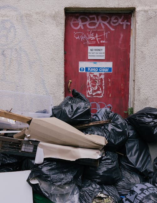 A pile of black rubbish bags and discarded cardboard boxes and packaging materials are stacked against a weathered red metal door embedded in a plain, off-white concrete wall with visible texture. The bags appear full and are loosely tied, with some of the packaging partially crushed or slightly torn. The door features multiple graffiti tags in white and light pink spray paint, overlapping each other, and displays signs including a white sign with black text stating, 'NO MONEY. KEEP ON TOYING. PRIMARIES OVERDRAFT,' as well as a blue and white sticker with the message, 'Keep clear.' The surrounding environment suggests an urban setting, possibly an alleyway or side entrance to a building, with no visible activity or people present. The lighting is soft and diffuse, typical of an overcast day, highlighting the textures of the materials and minor plant growth at the base of the wall. This scene demonstrates an instance of private waste accumulation, such as might be managed by a rubbish removal service, consistent with on-site clearance or alternative disposal options outside local authority collection systems, naturally supporting the context of rubbish removal in Crystal Palace.