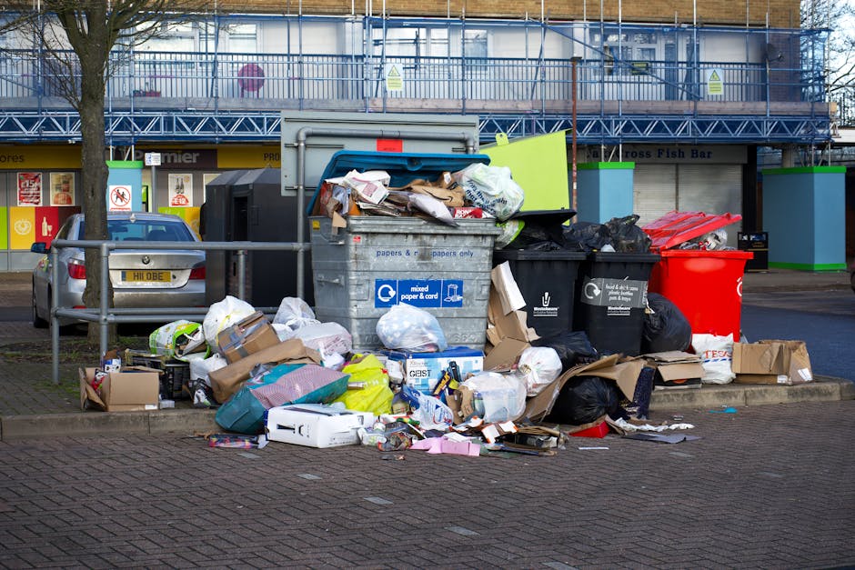 A large outdoor collection of mixed waste and rubbish overflowing from several black and grey recycling bins, with some items like cardboard boxes, paper, plastic bags, and packaging materials spilling onto the paved sidewalk area. The bins are placed near a metal railing and a parked silver car with a yellow license plate, on a brick-paved surface. In the background, there is a building under construction covered with blue scaffolding, and some shop fronts with signs indicating food establishments. The scene appears to be a result of an unauthorised or neglected rubbish accumulation, highlighting a need for proper waste removal services, such as those offered by independent rubbish collection companies like rubbishremovalcrystalpalace.com, to handle private waste disposal outside of official council services.