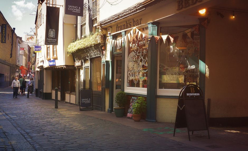 A narrow pedestrian street in an urban area with cobblestone paving, lined with small shops and restaurants. On the right, a building with a pale yellow exterior features large display windows decorated with festive bunting and interior artwork visible through the glass. In front of this shop, there are potted plants and a chalkboard sign with a logo for Bedford's. To the left, a series of storefronts and signage extend along the street, including a sign for Savino & Coombes and other business signs. Several pedestrians are seen walking along the street, some engaged in conversation, with light from the late afternoon sun casting soft shadows across the scene. The overall atmosphere suggests a lively commercial area, where independent shops and eateries may benefit from private waste handling, such as rubbish removal services provided by local companies, to maintain cleanliness in this busy setting.