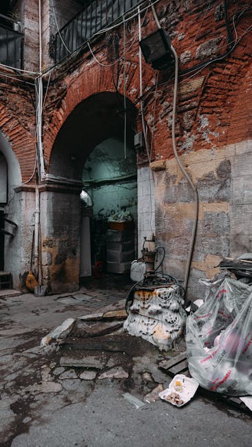 The image depicts an exterior scene featuring a brick archway entrance to a building, with reddish-brown and weathered bricks showing signs of age and wear. Above the arch, there are several exposed electrical wires and cables, some loose and hanging, along with a black external light fixture mounted to the brick wall. To the right of the arched doorway, there are piles of discarded debris, including crumpled plastic bags filled with rubbish and a large, partially deflated plastic bag leaning against the wall. On the ground, there is a small, rusted metal container or drum with a weathered surface, surrounded by bits of broken debris and loose gravel. The area appears to be a narrow alley or service entrance, with a concrete or paved surface that shows stains and cracks. The overall scene conveys an environment where private waste handling or rubbish clearance services, such as those provided by [COMPANY_NAME], might be involved in clearing clutter from the site or managing waste removal outside the building interior.