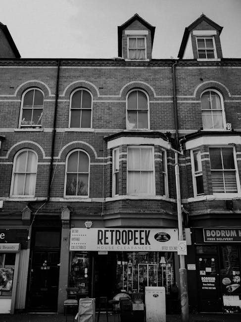 A black and white photograph of a multi-storey brick building with three upper windows featuring white trim and arched brickwork above them, situated on a street with retail shops on the ground floor. The storefront in the center displays a sign for 'Retropeek House Clearances,' indicating it provides private waste disposal or rubbish removal services. In front of the shop, several bags of waste or discarded materials are stacked on the pavement, along with a white bin and some smaller items. To the right, another shop sign for 'Bodrum' is partially visible. The building's upper architecture includes dormer windows with gabled roofs, contrasting with the lower shop windows and signage. The scene likely captures an urban environment where independent rubbish collection or house clearance services are offered, with visible waste materials set outside for removal, reflecting a typical setting for private rubbish removal in a residential or commercial area.